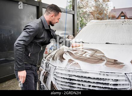 Young man washing car on carwash station outdoor. Handsome worker cleaning automobile, using sponge and soap. Stock Photo