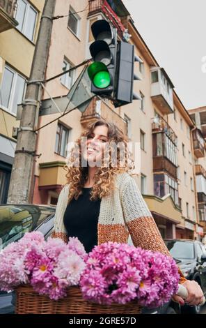 Close up of smiling cute young woman with curly hair riding bicycle with bouquet fresh pink flowers in centre city on traffic light background. Concept of walk on bicycle on city. Stock Photo