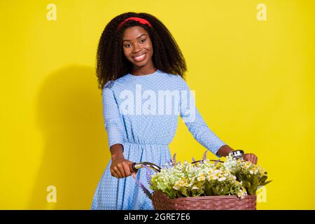 Photo portrait of curly girl wearing headband dotted blue dress riding bike with flowers basket isolated vivid yellow color background Stock Photo