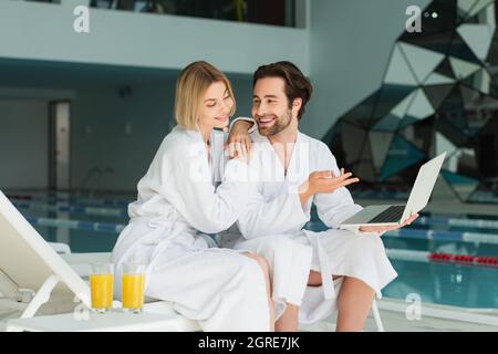 Smiling man pointing at laptop near girlfriend in bathrobe and orange juice in spa center Stock Photo