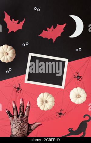 Abstract Halloween composition in red, black and white. Copy-space on blackboard. Paper silhouettes of Moon, bats, ghosts, eyes. Hand supports pyramid Stock Photo