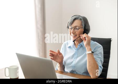 Grey-headed mature senior female office worker sitting at the desk with a laptop, wearing glasses, headset, and Fitbit, surfing the internet, writing an email, communicating with customers Stock Photo