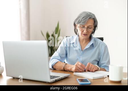 Serious mature senior female office worker business lady listening to an online webinar on laptop, taking notes in notebook, working on freelance project at home, remote work, learning at home concept Stock Photo