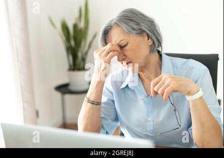 Tired and overworked stressed mature woman working on laptop at home indoor, female teacher feeling exhausted after lecturing students online, touching eyes. Smart working senior people and technology Stock Photo