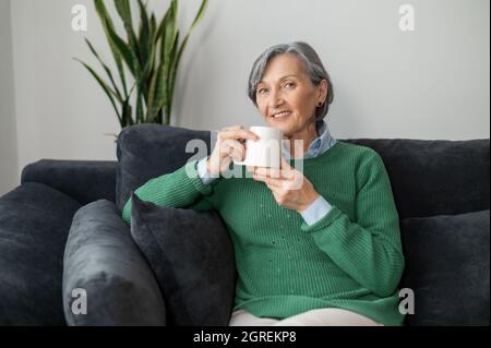 Charming senior mature lady holding mug of hot drink, looking at the camera, daydreaming and holding coffee, sitting on the couch in a cozy living room, enjoying free time