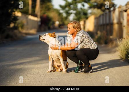 Woman crouching in an unpaved road next to a dog that sits distracted Stock Photo