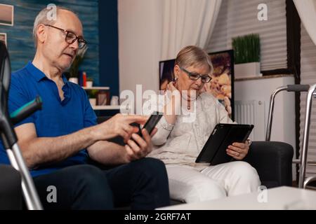 Old married people using digital devices and sitting together in living room. Senior woman looking at tablet while elder man holding smartphone on couch at home. Modern retired couple Stock Photo