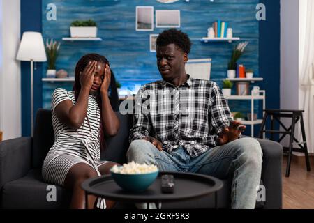 African american people watching scary movie on television and being afraid on sofa. Black woman covering eyes at TV while man looking at camera. Couple enjoying horror movie experience Stock Photo