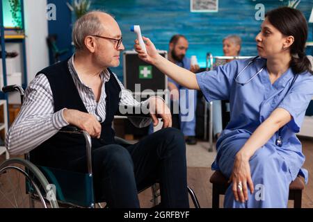 Support assistant worker measuring disabled senior man temperature using medical infrared thermometer during medicine therapy. Social services nursing elderly retired male. Healthcare assistance Stock Photo
