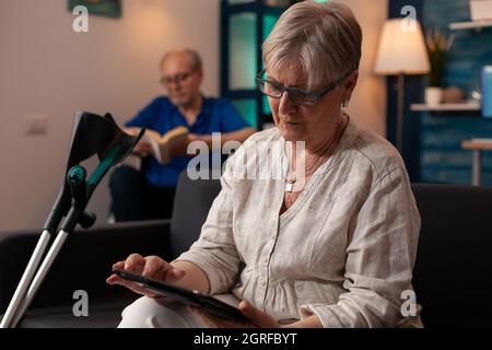 Caucasian old woman holding modern tablet on sofa with crutches while elder disabled man sitting in wheelchair with book in living room. Senior married couple enjoying retirement at home Stock Photo