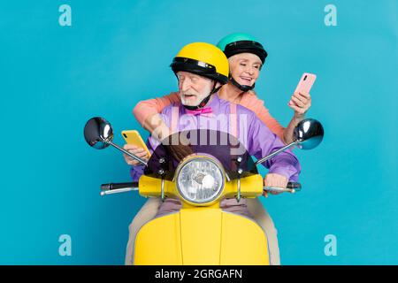 Portrait of two cheerful elderly retired pensioner friends riding bike using device gadget app isolated over bright blue color background Stock Photo