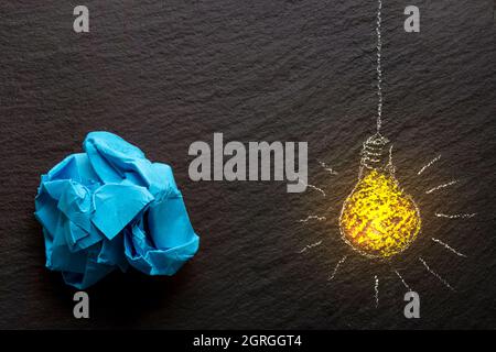 Chalk drawing of a yellow light bulb , lit, on  black rock texture, and a  crumpled blue piece of paper Stock Photo