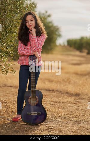 Pensive girl leaning on her guitar in a field of olive trees at sunset Stock Photo