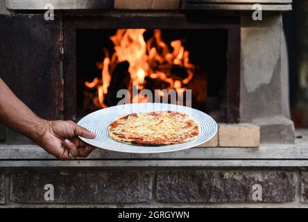 Arm holding a fresh baked pizza in front of a wood fired pizza oven. Stock Photo