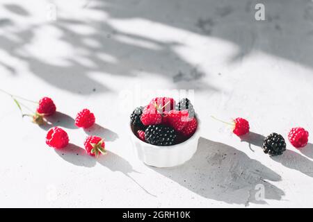 Bowl with raspberries and blackberries on gray concrete table with leaves shadow Stock Photo