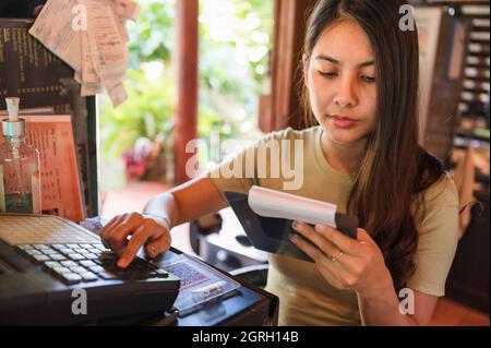 Young asian woman cashier checking order customer and pressing cash register at wooden counter in restaurant Stock Photo