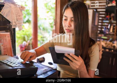 Young asian woman cashier checking order customer and pressing cash register at wooden counter in restaurant Stock Photo