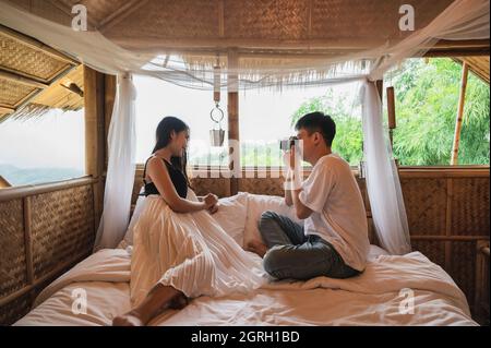 Young asian couple take photo with retro camera on the bed in wooden hut resort among the nature on vacation Stock Photo