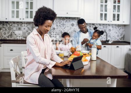 Focused woman working on digital tablet on bright kitchen while her husband feeding two daughters on background. African american family at home. Technology for lifestyles. Stock Photo
