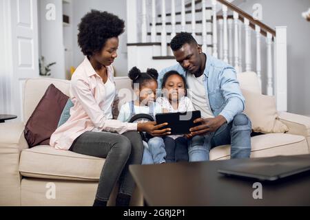 African american parents with two pretty daughters sitting on couch and watching cartoon on digital tablet. Positive family using modern gadget during leisure time at home. Stock Photo