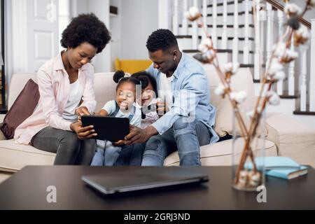 African parents and their two cute daughters watching cartoons on digital tablet while resting together on comfy couch. Concept of family, technology and leisure time. Stock Photo