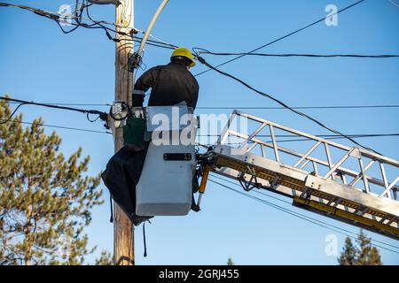 An upright shot of a technician from electric supply company, wearing a safety kit, sitting on the top of a ladder attached with the electricity pole Stock Photo