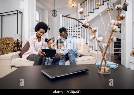 African parents and their two cute daughters watching cartoons on digital tablet while resting together on comfy couch. Concept of family, technology and leisure time. Stock Photo