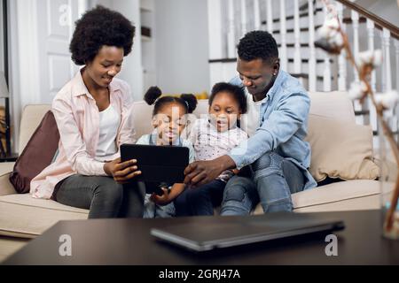 Happy african parents sitting on couch with two pretty daughters and using digital tablet for watching cartoons. Concept of family, leisure time and modern gadgets. Stock Photo