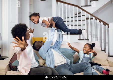 African mother, father and two daughters playing together while resting on comfy couch. Happy family spending time at home with fun and pleasure. Stock Photo
