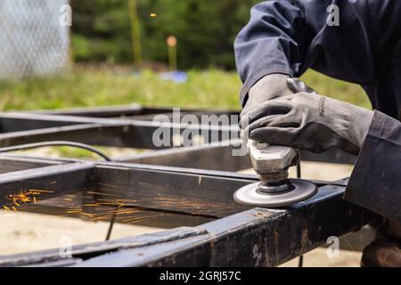 An image of a woman welder wearing protective gloves, running welding grinder on a metal surface, placed in a grassy field, in the daylight.  Stock Photo