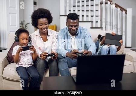 African wife and husband sitting on couch and playing video games on laptop with joystick. Two cute daughters sitting near with tablet and smartphone in hands. Modern technology for lifestyles. Stock Photo