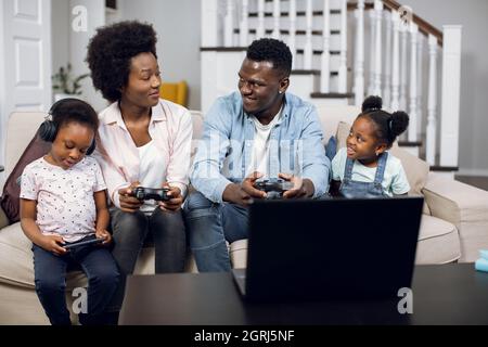 Smiling african man and woman sitting on sofa with wireless joysticks in hands and playing video games on laptop. Two cute daughters using smartphone and digital tablet. Stock Photo