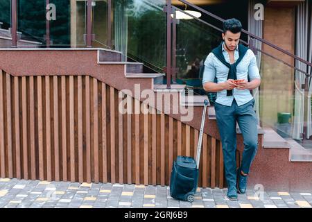 Attractive young Arabic man standing with suitcase in hand and posing for on camera, holding the phone and talking on it, put one foot before the Stock Photo