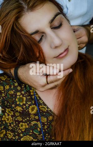 ortrait of a young red-haired woman with her mate's hands caressing her face while she has her eyes closed Stock Photo