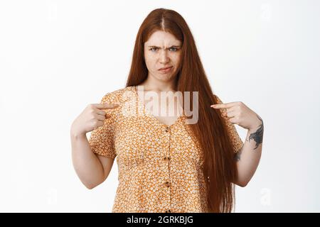 Image of angry and disappointed redhead girl, pointing at herself while sulking upset, standing over white background Stock Photo