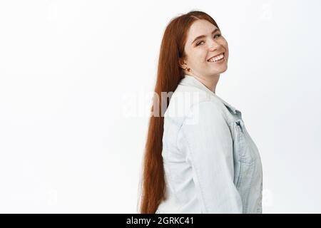 Portrait of young ambitious redhead girl, turn head at camera and smiling confident, standing in casual autumn clothes, white background Stock Photo