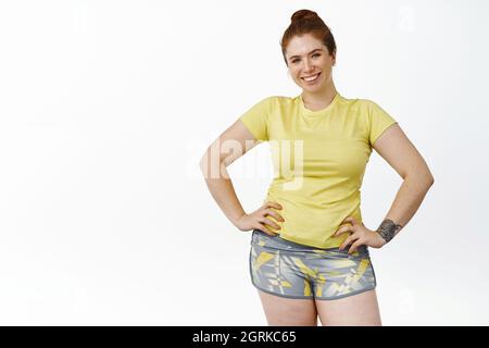 Portrait of young chubby woman in fitness clothing, workout in gym, smiling and looking enthusiastic at camera, white background Stock Photo