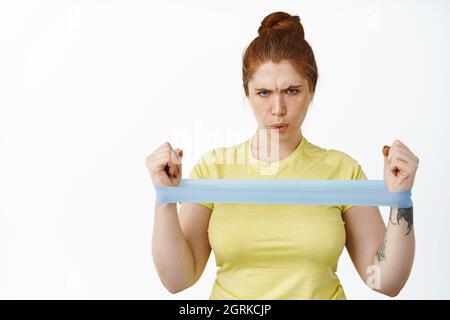 Serious redhead curvy woman doing workout with resitance band, fitness exercises on arms, stretching elastic rope, standing over white background Stock Photo