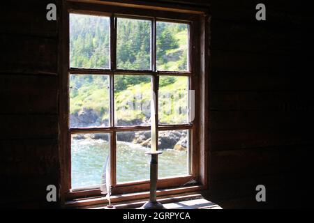 A candle in an antique candle holder and a quill pen in an ink well, on an old wooden desk in front of a window. View of coastline through window. Stock Photo