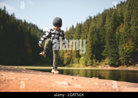 Small kid in a plaid shirt and gray hat on the forest lake coast. Childhood with nature loving concept Stock Photo