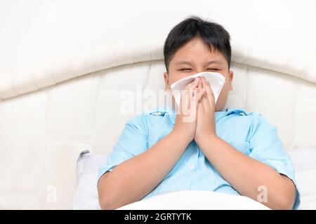 Sick Child. Asian Fat Boy Has Runny Nose And Blows Nose Into Tissue Sitting On Bed,