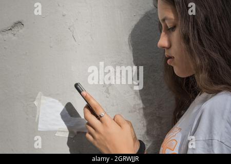 Side view of a teenage girl using her smartphone. She is texting while standing next to an old wall. The shadow of the girl's profile in the backgroun Stock Photo