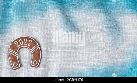 Ornate New year horseshoe for good luck in 2022 on blue white wavy fabric background. Beautiful cookie in horse shoe shape on wrinkled canvas texture. Stock Photo