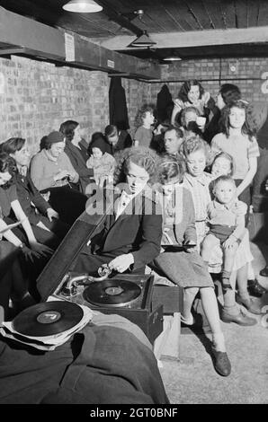 A young woman plays a gramophone in an air raid shelter in north London during 1940. A young woman places the gramophone needle on a record to bring some light relief to an air raid shelter, somewhere in north London. The rest of the shelterers appear to be enjoying her choice of music. In the background, one woman can be seen knitting, as others chat to pass the time. Stock Photo