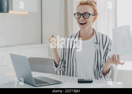 Overjoyed ginger woman holds papers works on project statistics celebrates finishing of work Stock Photo