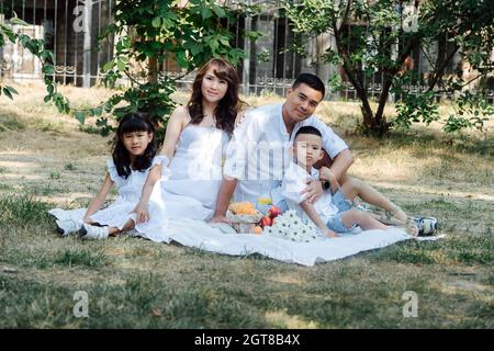 Mid distance portrait of an asian family in white clothes having picnic in a park, enjoying last warm days of the early fall. Parents and their childr Stock Photo