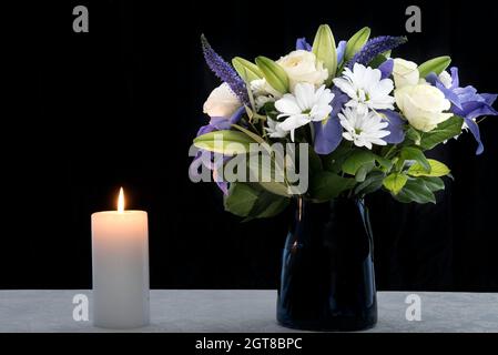 Funeral Bouquet purple White flowers and burning white candle, Sympathy and Condolence Concept on black background with copy space. Stock Photo