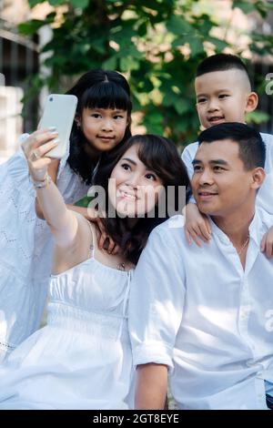 Beautiful asian family in white clothes taking selfie in a park. They are having picnic, enjoying last warm days of the early fall. Parents and their Stock Photo