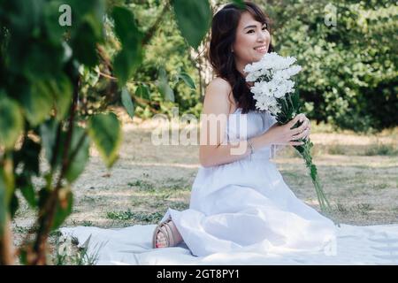 Smiling asian woman sitting on a blanket in a park, holding bouquet of white flowers She's wearing white dress, having picnic in a park, enjoying last Stock Photo