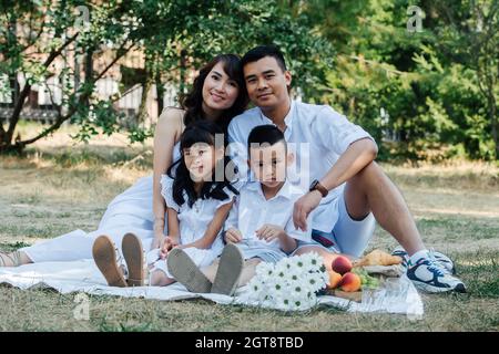 Family portrait of an asian family in white clothes having picnic in a park, enjoying last warm days of the early fall. Parents and their children in Stock Photo